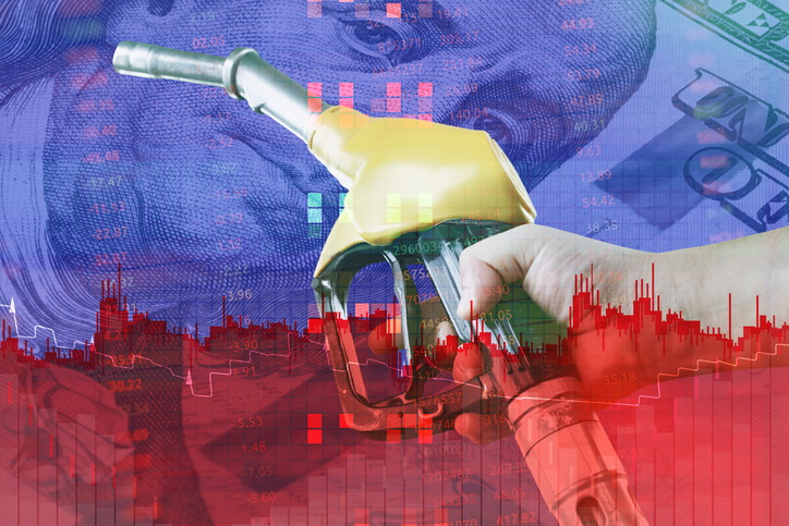 Hand holding a yellow gas pump nozzle over a background of US dollar bill and falling stock market charts. Concept rising fuel costs, energy industry crisis, economic recession, and oil prices. iStock